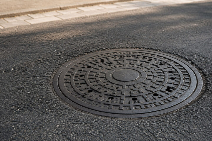 Warum sind Kanaldeckel rund? Runder Kanaldeckel auf einer Asphaltstraße aus schräger Perspektive